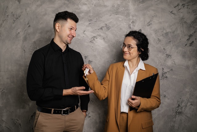 Person in mustard blazer and glasses handing a set of keys to person in black button-down